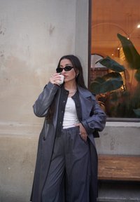 Longue veste en cuir grise portée sur un haut blanc, pantalon gris à fines rayures, et lunettes de soleil noires. Tenant une tasse blanche, avec de la verdure en arrière-plan.