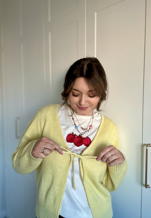Gul strikket cardigan med knyteåpning i livet, båret over en hvit t-skjorte med røde jordbær grafikk, sammen med en flerfarget halskjede.