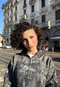 Kiharatukkainen nainen, joka käyttää kuvioitua vetoketjullista hupparia, seisoo aurinkoisella kaupunkikadulla, taustalla historiallisia rakennuksia ja polkupyöriä.