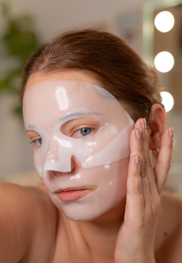 Jeune femme aux yeux bleus appliquant un masque facial translucide dans un intérieur doucement éclairé avec des lumières d'arrière-plan floues.