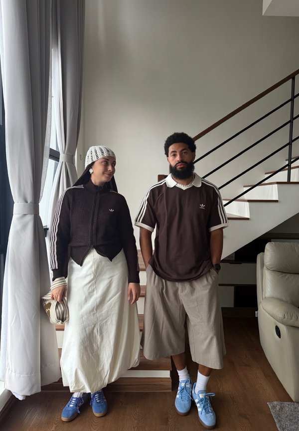 Dwie osoby stoją w pomieszczeniu przy schodach i oknie; obie mają na sobie brązowe bluzki Adidas, jasne spodnie i niebieskie trampki.