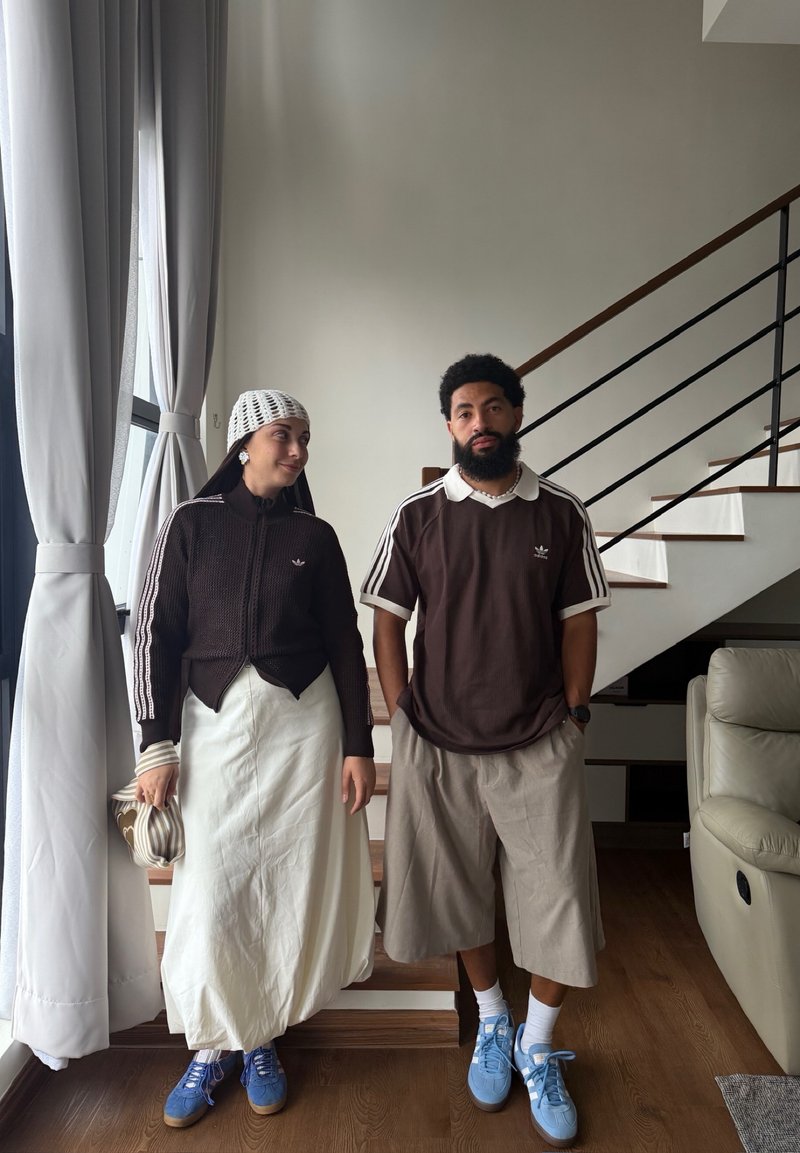 Deux personnes se tiennent à l'intérieur près d'un escalier et d'une fenêtre ; toutes deux portent des hauts Adidas marron, des bas clairs et des baskets bleues.