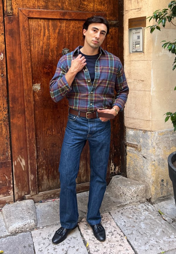 Un hombre con camisa de cuadros, vaqueros y zapatos negros posa delante de una gran puerta de madera mientras se sujeta el cuello de la camisa.