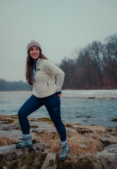 Joven mujer con ropa de invierno y botas de senderismo de pie sobre rocas cerca de un río, sonriendo y mirando hacia un lado, con árboles desnudos al fondo.