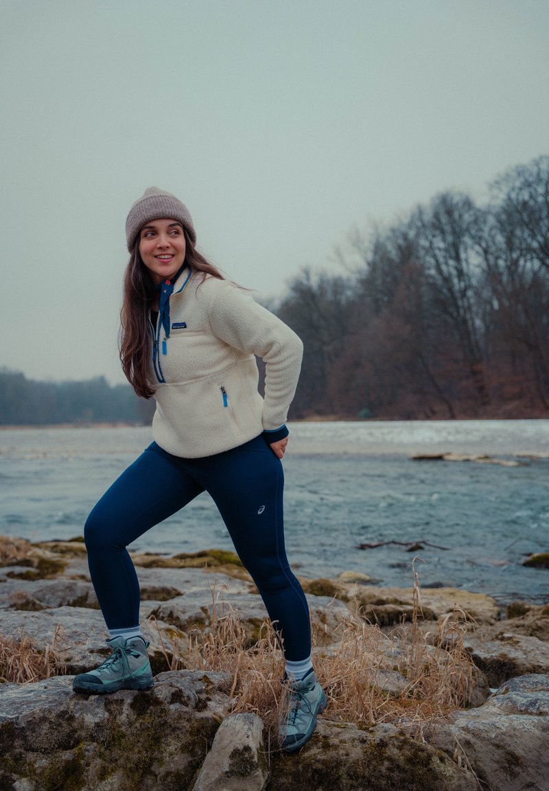 Jeune femme en vêtements d'hiver et bottes de randonnée debout sur des rochers près d'une rivière, souriante et regardant sur le côté, avec des arbres nus en arrière-plan.