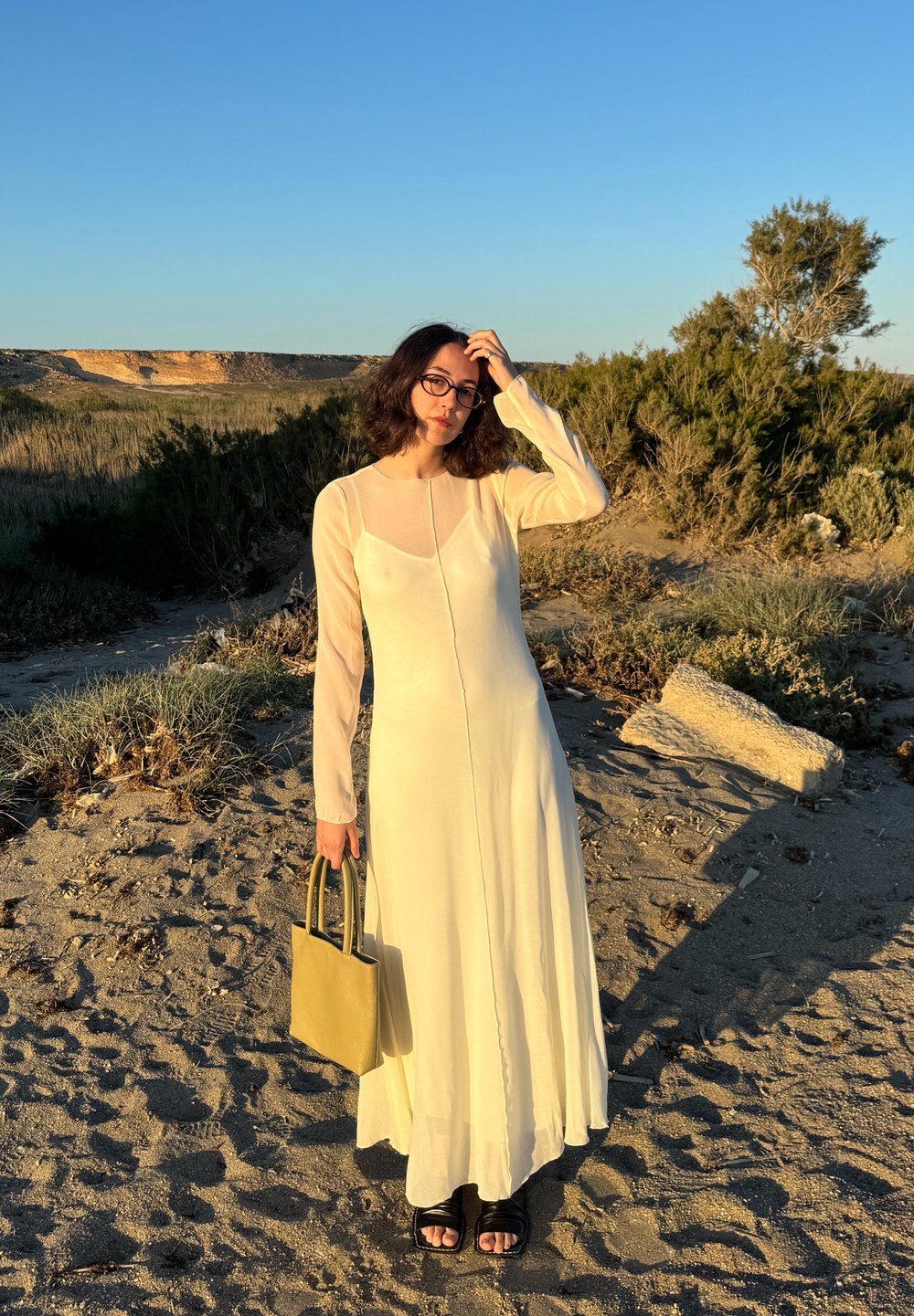 Rochie lungă, din material ivoire, cu mâneci lungi și decolteu rotund; asortată cu o geantă de umăr mică, de culoare verde măslin, și sandale negre.