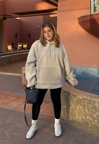 Kremfarget fleece-hettegenser med frontlomme og snøring, kombinert med svarte leggings, hvite sneakers og en liten marineblå veske.