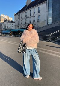 Jeune personne debout avec confiance, portant une veste rose duveteuse, un jean à jambes larges, des lunettes de soleil roses, et tenant un sac noir à pois blancs en plein air.