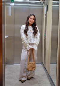 Jeune femme aux longs cheveux bruns portant un chemisier blanc et un pantalon, tenant un sac tissé, debout dans un ascenseur en acier inoxydable.