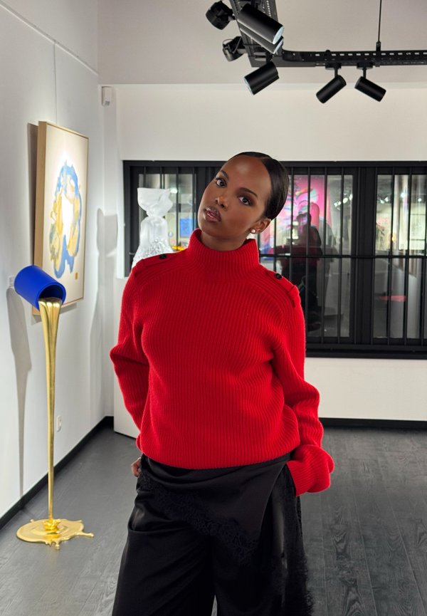 Donna con maglione a collo alto rosso e pantaloni neri in piedi in una galleria moderna con arte astratta e una scultura alle sue spalle.