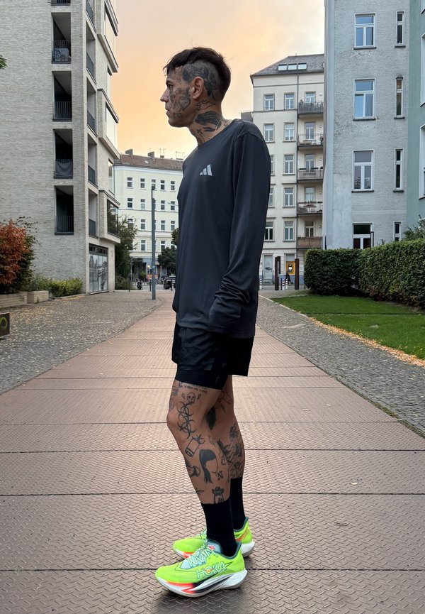 Joven con muchos tatuajes, vestido con ropa deportiva negra y zapatillas de correr de un verde brillante, está de pie en un camino urbano al atardecer.
