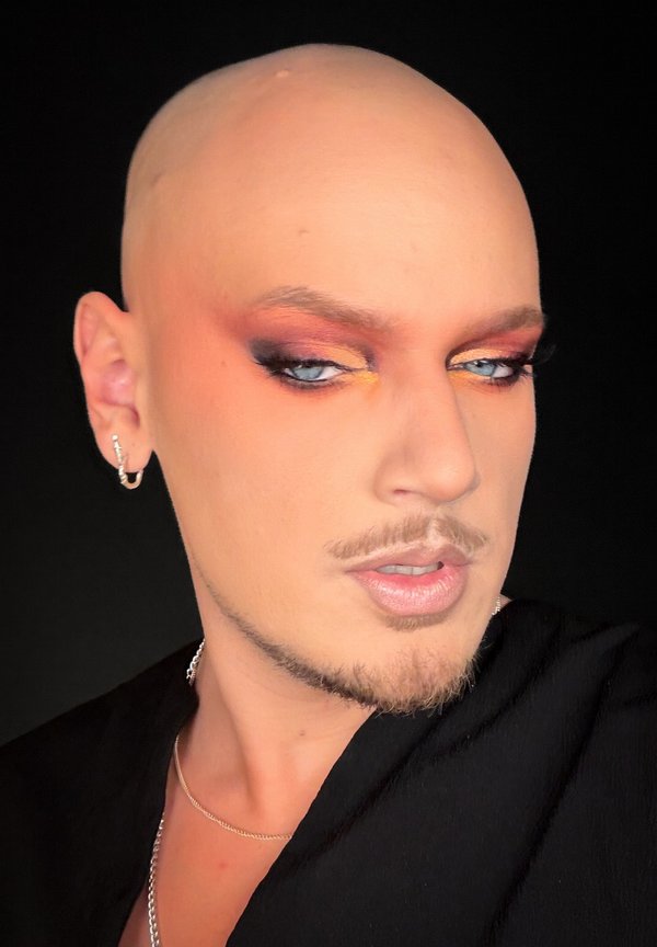 Skaldet person med stilfuld makeup i varme toner, blå øjne, trimmet skæg, iført øreringe og sort top mod en sort baggrund.