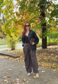 Melna ādas jaka virs melna mežģīņu blūzes, pelēki platās kājas bikses un melnbalti svītraini zābaciņi, turēdama brūnu maku.