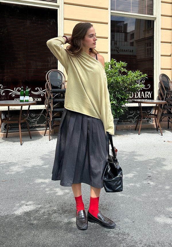 Femme avec un haut jaune clair à épaules dénudées, une jupe plissée noire, des chaussettes rouges, des chaussures noires et un sac noir, debout devant un café avec des tables et des chaises.