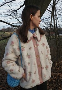 Veste en polaire crème à motifs floraux avec un devant en velours côtelé rose et un col à franges ; assortie à un sac bleu en crochet.