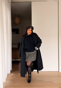 Femme portant un béret noir, un long manteau noir, un bracelet en perles, une jupe plissée grise et des bottes noires posant à l'intérieur sur un sol en bois.