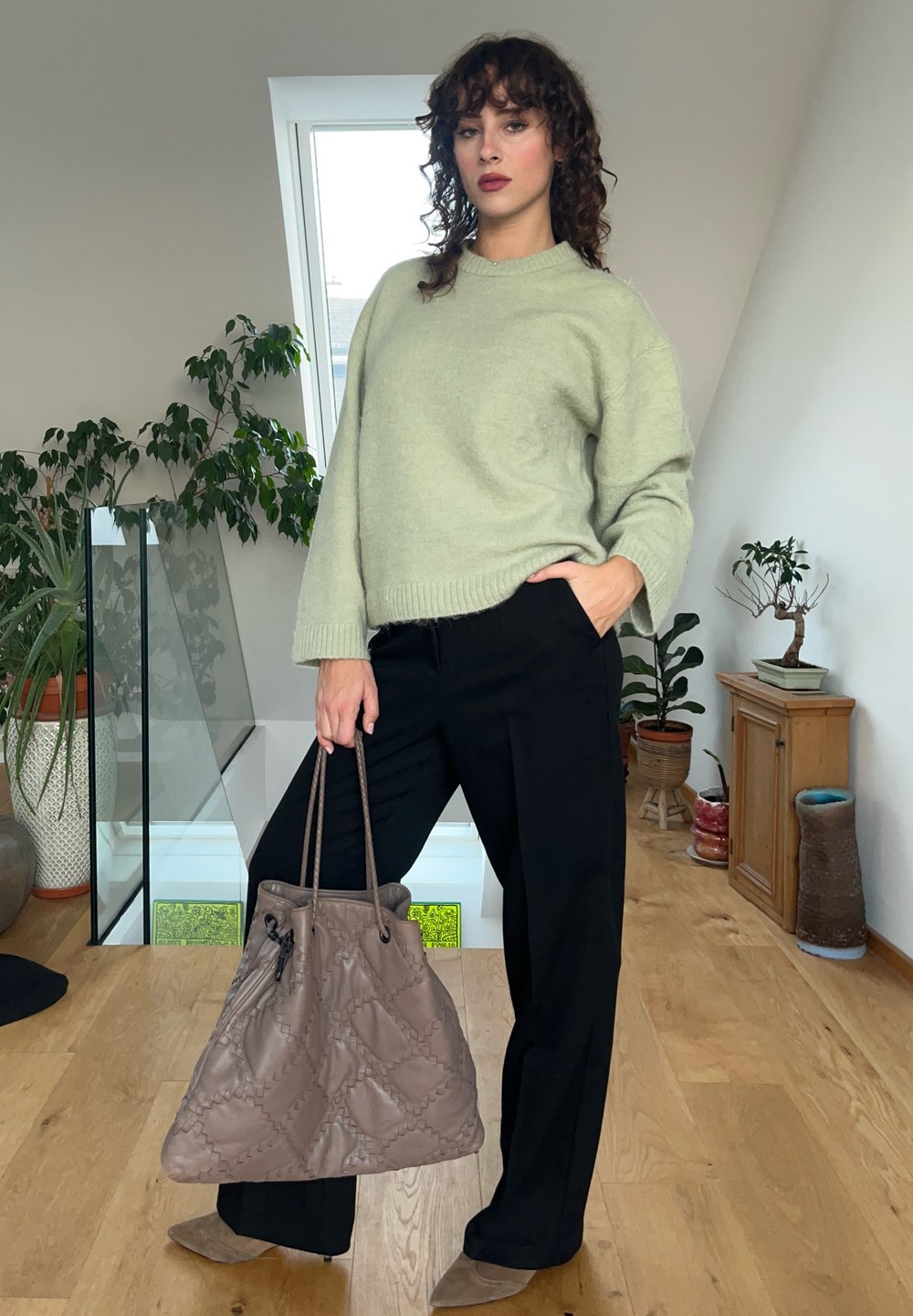 Ανοιχτό πράσινο πουλόβερ με ριγωτό στρίφωμα, σε χαλαρή εφαρμογή, συνδυασμένο με μαύρο παντελόνι με φαρδιές γραμμές. Κρατώντας μια απαλή ταμπά κούπα τσάντα με πλισέ.