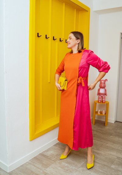 Mujer con vestido naranja y rosa, zapatos amarillos y bolso, de pie junto a un perchero amarillo montado en la pared en una habitación moderna.