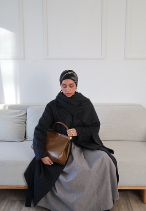 Bolso de cuero marrón con asa superior, llevado con un abrigo negro a rayas y una falda gris. El fondo muestra un sofá de color claro.