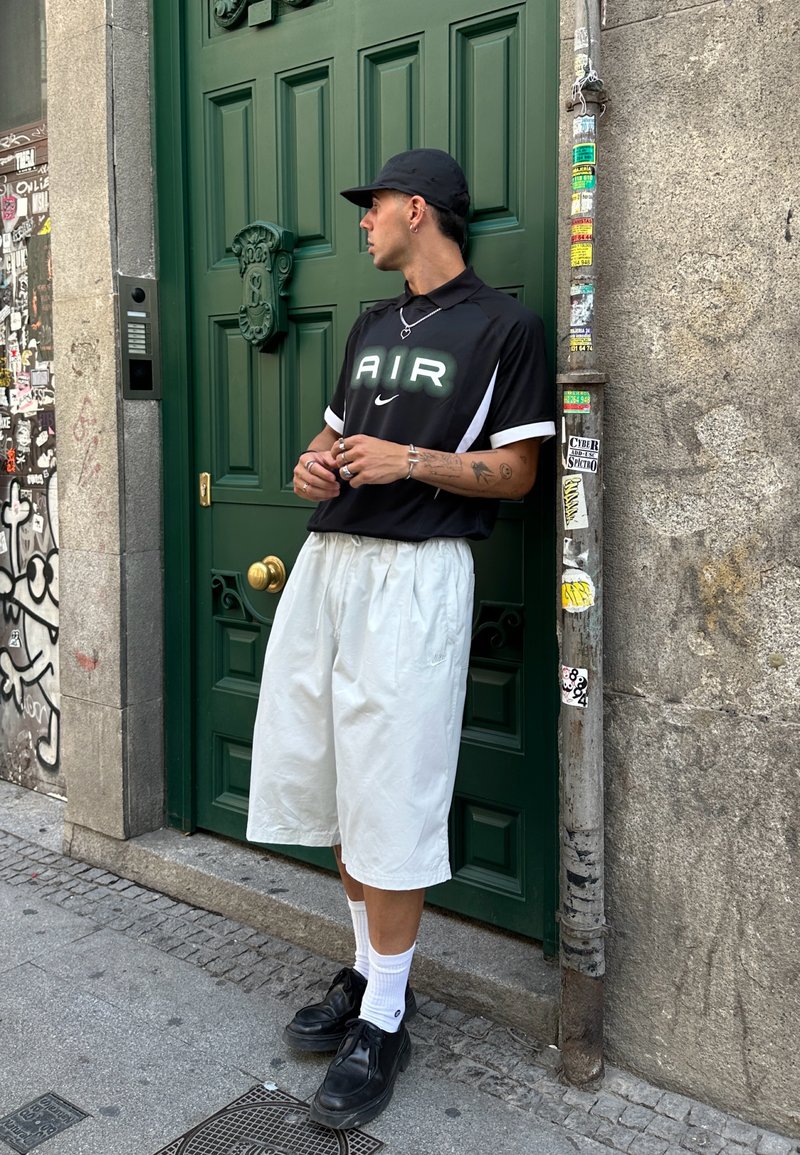 T-shirt de sport noir avec le logo "AIR", accents blancs, short blanc, chaussures noires, debout près d'une porte verte avec des motifs sur le mur d'arrière-plan.