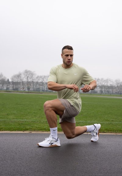 Atleta masculino con una camiseta Nike verde claro y pantalones cortos grises, realizando una zancada en una pista. Zapatillas blancas con detalles oscuros. Césped al fondo.