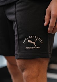 Czarne sportowe spodenki wykonane z bawełnianej tkaniny mieszanej, z białym wyszywanym logo "PUMA ATHLETICS" i napisem "założono w 1948 roku".
