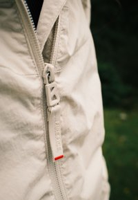 Šviesiai smėlio spalvos švarkas su matomu užtrauktuku. Užtrauktukas turi akivaizdžius metalinius elementus ir kilpą su užrašu "FIREICE", pažymėtą raudonu akcentu.