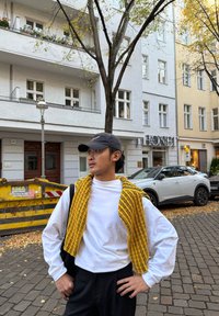 Witte lange mouw shirt gecombineerd met een geel gestreepte gebreide trui over de schouders gedrapeerd, zwarte broek en een zwarte pet, staande in de buitenlucht.