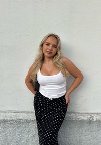 Hvit singlet, svart skjørt med hvite prikker, brun håndveske. Står mot en teksturert hvit vegg, med langt, bølgete blondt hår.