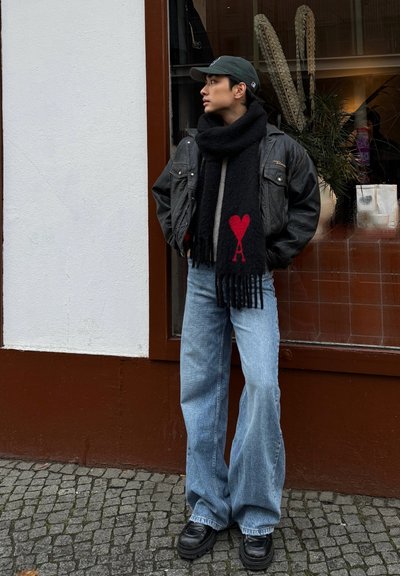 Chaqueta de cuero negra sobre un suéter gris, jeans azules de pierna ancha y una bufanda negra con diseño de corazón rojo y Torre Eiffel. Botas negras gruesas.