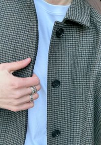 Manteau à motif pied-de-poule en gris et marron, avec un col, des boutons foncés, et une chemise blanche en dessous. Une main porte des bagues en argent.
