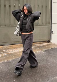 Sort bomberjakke med elastiske manchetter, sort langærmet top, løse sorte bukser med vide ben og chunky sorte sneakers, der holder en mønstret taske.