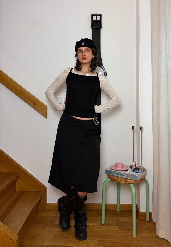 Joven mujer con falda y top negros posa con las manos en las caderas junto a un reloj de pared con forma de gran reloj negro cerca de unas escaleras de madera y una pequeña mesa.