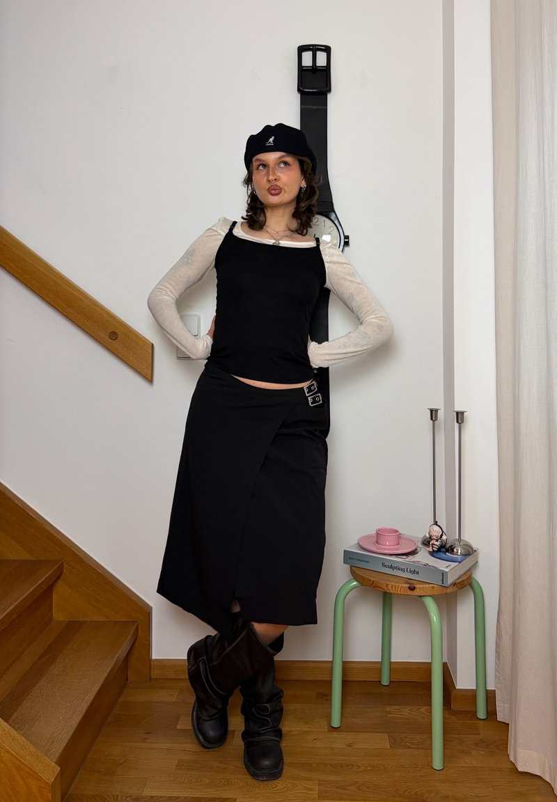 Joven mujer con falda y top negros posa con las manos en las caderas junto a un reloj de pared con forma de gran reloj negro cerca de unas escaleras de madera y una pequeña mesa.