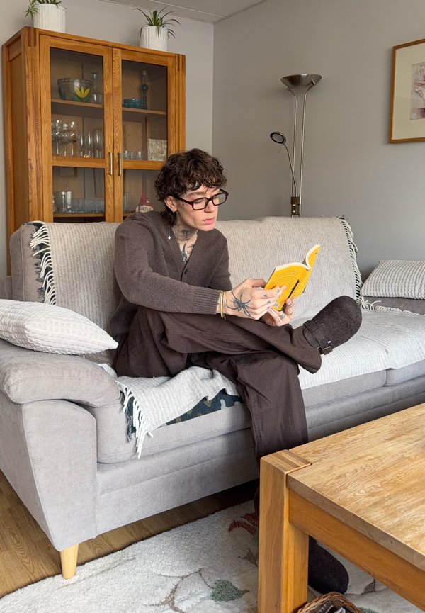 Person med briller og tatoveringer sidder med benene over kors på en grå sofa og læser en gul bog i en hyggelig stue.