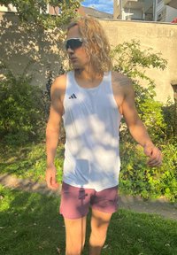 Conjunto atlético com uma camisa sem mangas branca com detalhes pretos e calções roxos. Cabelo loiro longo e ondulado e óculos de sol. Ambiente ao ar livre.