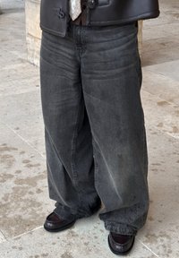 Laza szabású fekete farmer koptatott hatással, sötétbarna bőrcipőkkel, fényes felülettel. Alatta sík felület.