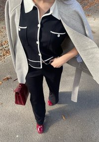 Μαύρη πλεκτή μπλούζα με λευκές λεπτομέρειες και τσέπες, συνδυασμένη με μαύρο παντελόνι. Γκρι παλτό ριγμένο στον ώμο, κρατώντας μια μπορντό τσάντα. Κόκκινα υποδήματα.