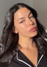 Muotokuva henkilöstä, jolla on pitkät, laineikkaat mustat hiukset ja kirkas ihonsävy. Hänellä on päällään musta yläosa, jossa on valkoisia yksityiskohtia, ja hänet valaisee luonnonvalo.