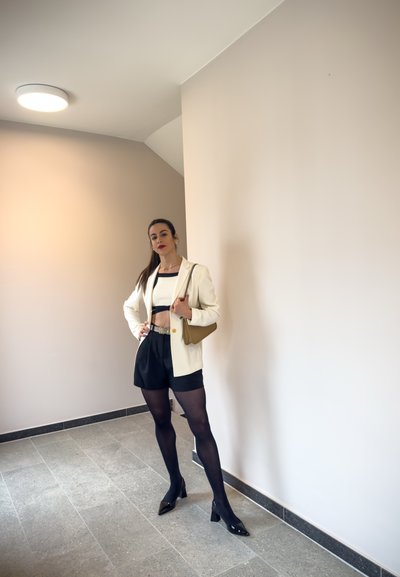 Blazer blanco sobre un top corto, pantalones cortos negros de cintura alta y tacones negros. Medias texturizadas; bolso pequeño marrón, de pie en un área bien iluminada.