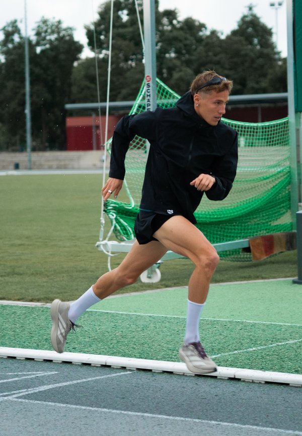 Mladić u sportskoj odjeći sprinta na vanjskoj stazi, u sredini koraka, s zelenom mrežom i drvećem u pozadini.