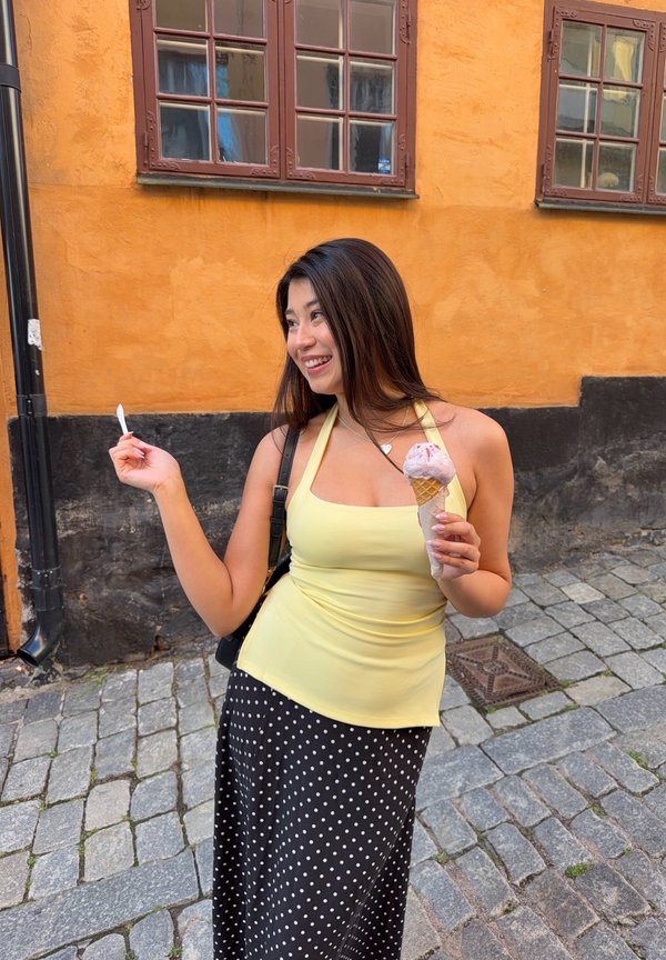 Femme souriante en haut jaune et jupe à pois tenant un cornet de glace et une cuillère dans une rue pavée avec un mur jaune et noir derrière.