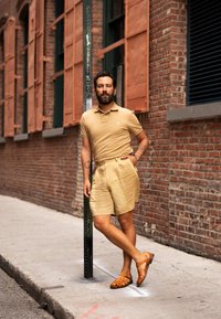 Tann polo t-shirt och matchande shorts i texturerat tyg. Bruna lädersandaler. Urban miljö med tegelvägg och grön stolpe.