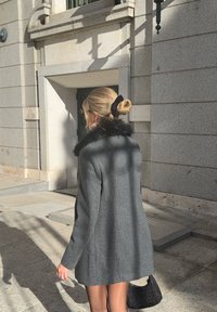 Szőke haját kontyba kötött nő, szürke kabátban fekete szőrme gallérral, fekete kézitáskával a kezében, napsütésben egy kőépület mellett sétál.