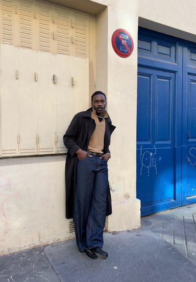 Hombre con abrigo negro, suéter marrón y jeans anchos apoyado contra una pared beige cerca de puertas azules y un cartel circular rojo de prohibido estacionar.