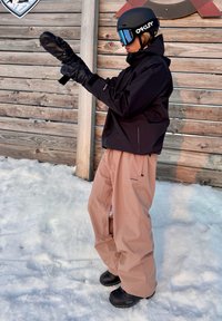 Črna nepremočljiva jakna, bež snežne hlače in črne čevlje. Velike črne mittens in zaščitna očala s modrim odtenkom dopolnjujejo zimski športni komplet.