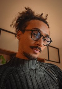Personne aux cheveux bouclés, portant des lunettes rondes noires et un pull en tricot gris foncé, regardant vers le bas à l'intérieur avec des œuvres d'art encadrées au mur derrière.