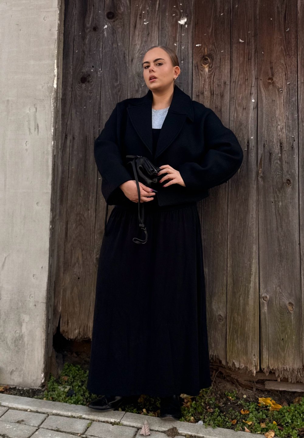 Zwarte cropped jas over een grijze top, gecombineerd met een lange zwarte rok. Het onderwerp houdt een kleine zwarte tas vast en staat tegen een houten achtergrond.