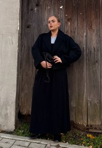 Zwarte cropped jas over een grijze top, gecombineerd met een lange zwarte rok. Het onderwerp houdt een kleine zwarte tas vast en staat tegen een houten achtergrond.