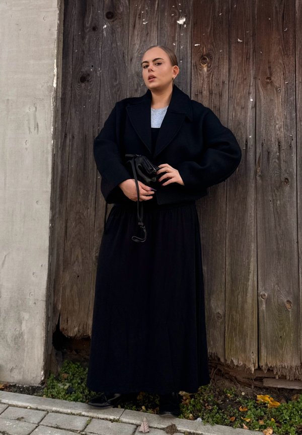 Musta lyhyt takki harmaan paidan päällä, yhdistettynä pitkään mustaan hameeseen. Aihe pitää pientä mustaa laukkua ja seisoo puista taustaa vasten.